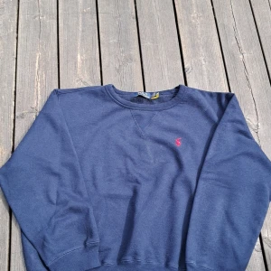 Blå sweatshirt från Polo Ralph Lauren - Mörkblå sweatshirt från Polo Ralph Lauren med klassisk rund hals och broderad röd logga på bröstet. Tröjan har långa ärmar och ribbade muddar vid ärmslut och nederkant. Perfekt för en avslappnad stil.