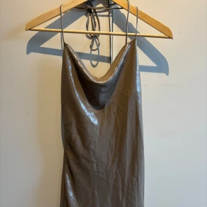 Brun glansig klänning med halterneck - Säljer en brun glansig klänning med halterneck och draperad urringning. Klänningen har smala axelband som knyts i nacken och en elegant, slank siluett. Perfekt för dig som vill ha en stilren och trendig look.