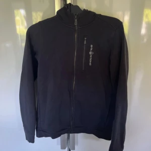 Svart hoodie med dragkedja från Sail Racing - Svart hoodie från Sail Racing med hel dragkedja framtill och hög krage. Jackan har en bröstficka med dragkedja och Sail Racing-logga på bröstet. Mjuk och bekväm, perfekt för kyligare dagar.