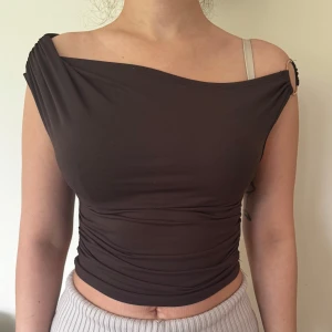 Brun oneshoulder topp med ringdetalj - Trendig brun oneshoulder topp med en snygg ringdetalj på axeln. Toppen är tight och croppad, perfekt för att sticka ut. Gjord i mjukt bomullsmaterial som sitter skönt mot kroppen. Finns även en likadan fast i rött