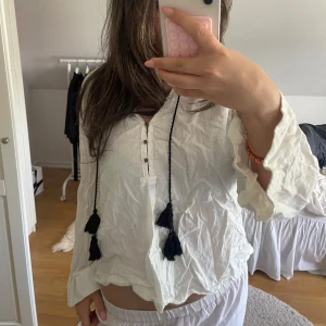 Vit blus med knappar och tofsar - Säljer en luftig vit blus med långa ärmar, knappar framtill och svarta tofsar som detalj. Blusen har en avslappnad passform och är perfekt för vår och sommar. Superfin att styla till både jeans och kjol! Strl: s/m