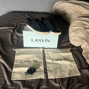 Svarta sneakers från Lanvin med vit sula - Säljer ett par svarta sneakers från Lanvin med vit platt sula och rund tå. Skorna har snörning och en glansig tådel i svart. Klassisk och stilren design, perfekt för dig som gillar lyxiga detaljer.