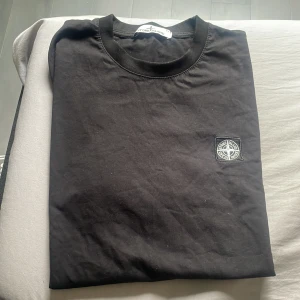 Svart t-shirt från Stone Island - Säljer en svart t-shirt från Stone Island med klassisk rund hals och logotyp-patch på bröstet. Enkel och stilren design som passar perfekt till jeans eller shorts. Obs dåliga bilder den ör helt svart