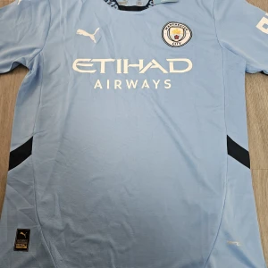 Manchester City hemmatröja Puma - Säljer en ljusblå Manchester City fotbollströja från Puma med klubbmärke och Etihad Airways-tryck på bröstet. Tröjan har korta ärmar, svart detalj på sidorna och mönstrad krage. Tillverkad i lätt och ventilerande material.