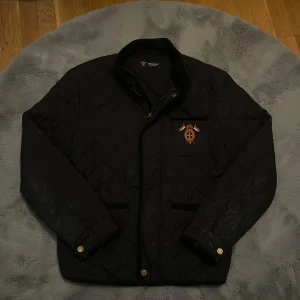 Mörkblå quiltad jacka från Morris - Säljer en mörkblå quiltad jacka från Morris med broderad detalj på bröstet och ståkrage i manchester. Jackan har dragkedja och knappar framtill samt fickor och justerbar midja. Perfekt för sena sommarkvällar. (Passar som M/L). 