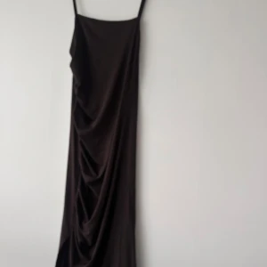 Svart långklänning med smala axelband - En stilren svart långklänning med smala axelband och rak siluett. Klänningen har en elegant drapering framtill och är tillverkad i ett mjukt, glansigt material som ger en lyxig känsla.