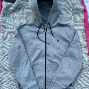 Ralph Lauren zip hoodie - Säljer en klassisk grå hoodie från Polo Ralph Lauren med dragkedja och svart broderad logga på bröstet. Tröjan har långa ärmar, huva med snörning och praktiska fickor framtill. Perfekt för en avslappnad stil.