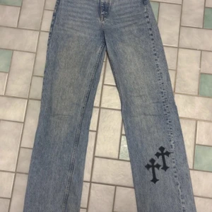 Blå jeans med svarta kors från Zara - Säljer ett par blå jeans från Zara med raka ben och två svarta kors som detalj på ena benet. Klassisk femficksmodell med knapp och dragkedja framtill. Perfekta för dig som vill sticka ut lite extra.