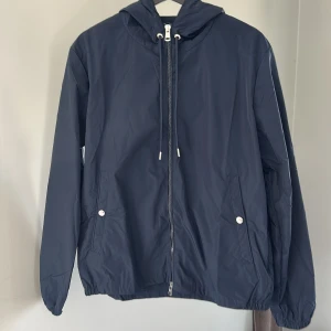 Mörkblå vindjacka från Moncler - Säljer en mörkblå vindjacka från Moncler med huva och dragkedja framtill. Jackan har vita och röda detaljer på huvan, två fickor med knappar och Moncler-logga på ärmen. Perfekt för blåsiga dagar.