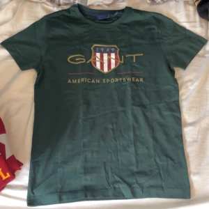 tartan green gant t-shirt - Säljer en grön t-shirt från Gant med tryckt logga och texten 'American Sportswear' på bröstet. Klassisk rund hals och normal passform. Perfekt för en avslappnad stil.