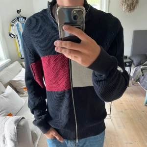Stickad ziptröja från Tommy Hilfiger - Säljer en stickad långärmad tröja från Tommy Hilfiger med dragkedja. Tröjan är mörkblå med breda block i rött och vitt framtill. Klassisk ståkrage och ribbade muddar. Sparsamt använd, fint skick.