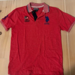 Röd pikétröja från U.S. Polo Assn. - Röd pikétröja med broderad blå USPA-logga och emblem på bröstet. Kragen är randig i rött, vitt och blått, och tröjan har knappar vid halsen. Perfekt för en sportig och stilren look.