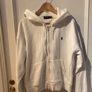 Vit hoodie med dragkedja från Polo Ralph Lauren - Vit hoodie från Polo Ralph Lauren med dragkedja framtill och två fickor. Klassisk design med broderad logga på bröstet och justerbar huva med snören. Perfekt för en avslappnad stil. XL barnstorlek, som en S.