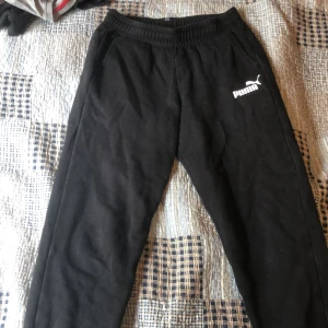 Svarta mjukisbyxor från Puma - Svarta mjukisbyxor från Puma med resår i midjan och vit Puma-logga på vänster lår. Perfekta för chill eller träning, mjukt material och klassisk passform.
