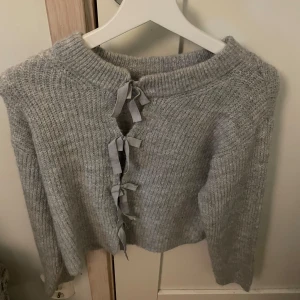 Grå stickad kofta med rosett från Gina Tricot - Superfin grå stickad kofta från Gina Tricot med långa ärmar och dekorativa rosetter framtill. Koftan är croppad och har en mysig, mjuk känsla. Perfekt för lager-på-lager och enkel att styla till olika outfits.(aldrig använd)