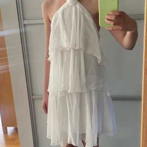 Vit volangklänning med rosettdetalj - Säljer en vit kortklänning med flera lager volanger och en snygg rosettdetalj framtill vid halsen. Klänningen är ärmlös, har öppen rygg och axelband som korsas bak. Perfekt för sommarens tillfällen!
