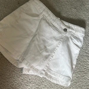 Vita omlottshorts i jeans - Säljer ett par vita omlottshorts i jeansmaterial med fickor fram och bak samt knapp i midjan. Snygg och unik omlottdesign som ger en cool look. Perfekta för sommaren!