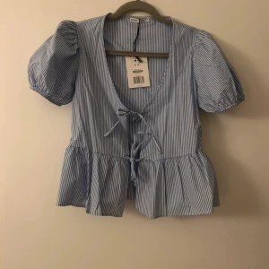 Blårandig blus med puffärm och knyt - Säljer en söt blårandig blus med puffärmar och knyt framtill. Blusen har v-ringning och volang nedtill, perfekt för en fräsch och somrig look.