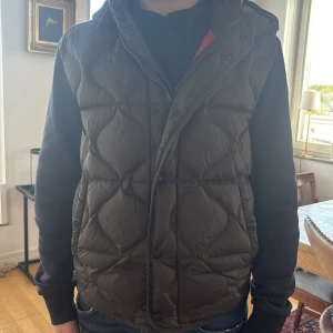 Mörk grön/även blå moncler cardigan - Säljer en mörkgrön/blå cardigan från Moncler med quiltat mönster och huva. Västen har knappar och dragkedja framtill samt Moncler-logga på ärmen. Perfekt för lager-på-lager och stilren look.