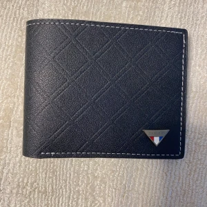 Svart plånbok  - Snygg svart plånbok i skinn med diskret rutmönster och vit söm runt kanterna. Plånboken har flera kortfack och ett fönster för ID-kort. Metallogo på framsidan. Perfekt för att hålla ordning på kort och kontanter.