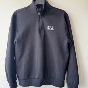Svart halvzip sweatshirt från Emporio Armani EA7 - Svart sweatshirt med halv dragkedja från Emporio Armani EA7. Tröjan har lång ärm, hög krage och en vit logga på bröstet. Knappt använd!