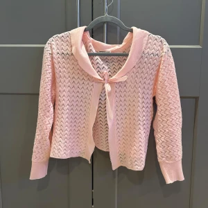 Ljusrosa stickad kofta med rosett från Courtelle - Supersöt ljusrosa stickad kofta från Courtelle med dekorativ rosett framtill. Koftan har ett spetsliknande mönster, långa ärmar och en mjuk krage. I strl S