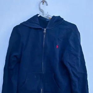 Mörkblå hoodie från Polo Ralph Lauren - Säljer en mörkblå hoodie från Polo Ralph Lauren med dragkedja och huva. Hoodien är från kids brand store och passar 165 personer. Tröjan har en liten röd broderad logga på bröstet och två fickor framtill. Perfekt för en avslappnad stil.