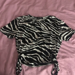 Zebramönstrad topp med knyt från Tally Weijl - Säljer en svartvit zebramönstrad topp från Tally Weijl i storlek S. Toppen har korta ärmar och knytband i sidorna nertill för en snygg detalj. Perfekt för dig som gillar djurmönster och vill sticka ut.