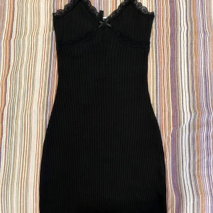 Svart ribbad klänning med spets från H&M Divided - Säljer en svart ribbad klänning från H&M Divided i storlek XXS. Klänningen har spetsdetaljer vid urringningen och en liten rosett framtill.