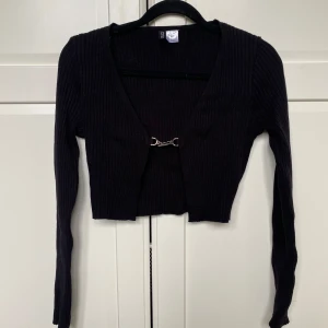 Svart croppad ribbad kofta från H&M - Säljer en svart croppad kofta från H&M  i ribbat material. Koftan har lång ärm, djup v-ringning och stängs framtill med en metallhake. Perfekt att styla över ett linne eller klänning.