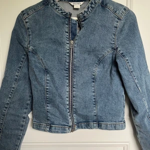 Blå jeansjacka från Monki - Säljer en ball klassisk blå jeansjacka från Monki med dragkedja framtill. Jackan har rundad halsringning och markerade sömmar för en snygg passform. Perfekt att slänga över en t-shirt eller hoodie.