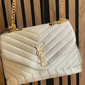Beige quiltad axelväska  - Supersnygg beige axelväska  i mjuk sammet med quiltad design och guldiga kedjor. Väskan har det ikonisk  framtill och en stilren, lyxig look som verkligen sticker ut. Perfekt storlek för det viktigaste och ger en exklusiv vibe till din outfit.
