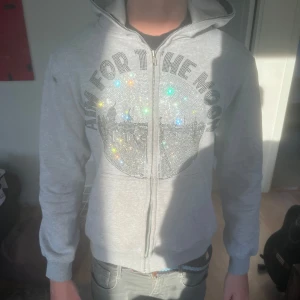 Grå hoodie med glittrig text - Snygg ljusgrå hoodie med dragkedja och huva. Framsidan har ett stort glittrigt tryck med texten 'AIM FOR THE MOON' och massor av små stenar som reflekterar ljuset. Perfekt för dig som vill sticka ut lite extra.