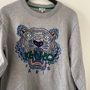 Grå sweatshirt med tigerbrodyr från Kenzo - Grå sweatshirt från Kenzo med ikoniskt broderat tigerhuvud i blått, grönt och beige framtill. Tröjan har rund halsringning, långa ärmar och en relaxed passform. Perfekt statement-plagg för dig som gillar streetstyle och coola detaljer. Nypris: 3999kr