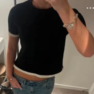 Svart t-shirt med vita detaljer från ZARA - Svart t-shirt från ZARA med vita ribbade kanter vid ärmslut och nederkant. Klassisk rund halsringning och rak passform. Perfekt till jeans eller kjol för en stilren look.