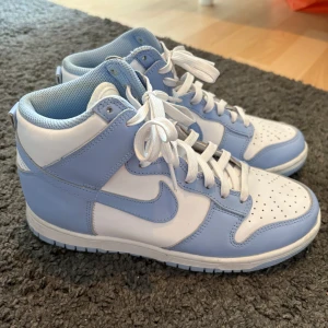 Nike Air Jordan vit/blå sneakers - Snygga Nike Dunk High sneakers i vitt och ljusblått skinn. Klassisk siluett med högt skaft, perforerad tå och vit snörning. Perfekta för dig som vill sticka ut med en fräsch och sportig look. Ikonisk Swoosh-logga på sidan och bekväm platt sula. Endast använda en gång 