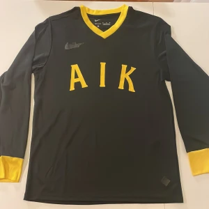 AIK långärmad fotbollströja Nike - AIK långärmad fotbollströja replika från Nike i svart med gula detaljer vid hals och ärmslut. Framsidan har stora gula bokstäver 'AIK' och diskret Nike-logga. Tillverkad i ett lätt och sportigt material, perfekt för match eller träning. Endast handtvätt gäller!