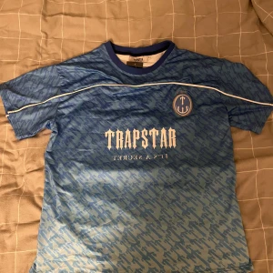 Blå Trapstar fotbollströja S - Helt ny aldrig använd, storlek S men passar som M. Snygg blå fotbollströja från Trapstar med vit logga och texten 'IT'S A SECRET' på bröstet. Tröjan har ett mönster över hela ytan, vita detaljer och nummer 22 på ryggen. Tillverkad i lätt och ventilerande material, perfekt för fotboll eller streetwear.
