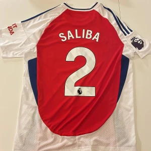 Arsenal Saliba #2 hemmatröja Adidas - Arsenal hemmatröja replika med Saliba och nummer 2 på ryggen. Adidas-logga, klubbmärke och Premier League-patch på ärmen. Röd front, vita ärmar och sidor med blå detaljer. Tillverkad i lätt och ventilerande material, perfekt för match eller träning. Endast handtvätt!