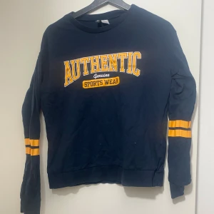 Mörkblå sweatshirt från DIVIDED S - Collegeinspirerad mörkblå sweatshirt från DIVIDED i storlek S. Tryck med texten 'AUTHENTIC SPORTS WEAR' i gult och vitt framtill samt gula ränder på ärmarna. Mjuk bomullskänsla och klassisk passform med rund hals och långa ärmar.