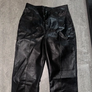 Svarta raka byxor i skinnimitation - Snygga svarta byxor i skinnimitation med raka ben och hög midja. Byxorna har fickor fram och bak samt markerade sömmar längs benen för en clean look. Perfekta för dig som vill ha en edgy vibe till outfiten.