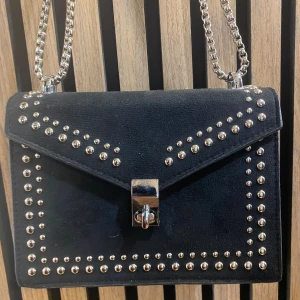 Svart mocka kuvertväska med nitar - Snygg svart kuvertväska i mocka med silverfärgade nitar längs kanterna och ett coolt lås framtill. Väskan har en kedjerem i silver som ger en edgy vibe. Perfekt för dig som vill sticka ut med detaljer och stilren design.