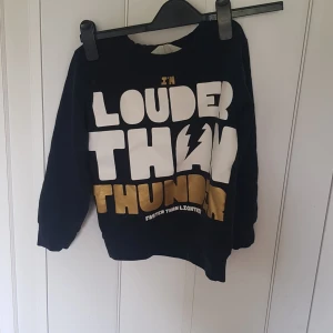 Svart sweatshirt med tryck från H&M - Cool svart sweatshirt från H&M med stort tryck i vitt och guld: 'I'M LOUDER THAN THUNDER' och blixtdetalj. Tröjan har långa ärmar och ribbade muddar. Perfekt för dig som gillar statement-plagg och vill sticka ut.