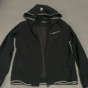 Svart softshelljacka Sail Racing S - Svart softshelljacka från Sail Racing med huva och vita detaljer längs kanterna. Jackan har dragkedja framtill, logga på bröstet och är tillverkad i stretchig polyesterblandning. Perfekt för dig som gillar sportig och clean stil.