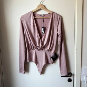 Ljusrosa omlott-body från NLY One - Supersnygg ljusrosa body från NLY One med omlott och djup v-ringning. Långärmad modell i glansigt, stretchigt material som sitter tajt och markerar midjan. Perfekt att styla med jeans eller kjol för en trendig look. Storlek XS. Helt ny med tags/ prislapp fortfarande kvar. 