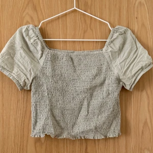 Grågrön blus från Kappahl - Fin grågrön blus från Kappahl med smockad kropp och puffiga korta ärmar. Toppen har fyrkantig ringning och elastiska detaljer som ger en skön passform. 