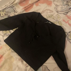 Svart ribbad blus från BIK BOK - Svart blus från BIK BOK med ribbat tyg och v-ringning. Modellen har långa ärmar och knappar framtill. Perfekt för dig som gillar en stilren och enkel look med lite edge.