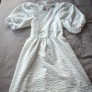 Vit kortklänning från Gina Tricot - Supersöt vit kortklänning från Gina Tricot i storlek XS. Klänningen har puffiga korta ärmar, strukturmönstrat tyg och knytning i nacken.