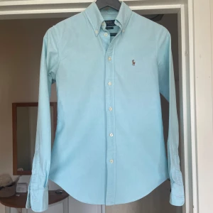 Ljusblå skjorta från Polo Ralph Lauren XS - Snygg ljusblå skjorta från Polo Ralph Lauren i custom fit. Klassisk button-down krage, broderad logga på bröstet och vita knappar. Skjortan är långärmad och tillverkad i mjuk bomull, perfekt för en clean och stilren look. OBS, finns en fläck på höger arm, men går bort i tvätten! Skjortan passar en som är mellan 160 cm till 170 cm, hör av er vid intresse! 
