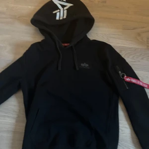 Svart hoodie Alpha Industries S - Svart hoodie från Alpha Industries med stor vit logga på ryggen och mindre logga på huvan. Klassisk känguruficka, snörning i huvan och ikonisk röd 'Remove Before Flight'-tag på ärmen. Mjuk bomullsblandning och dragkedjeficka på ärmen för extra attityd.